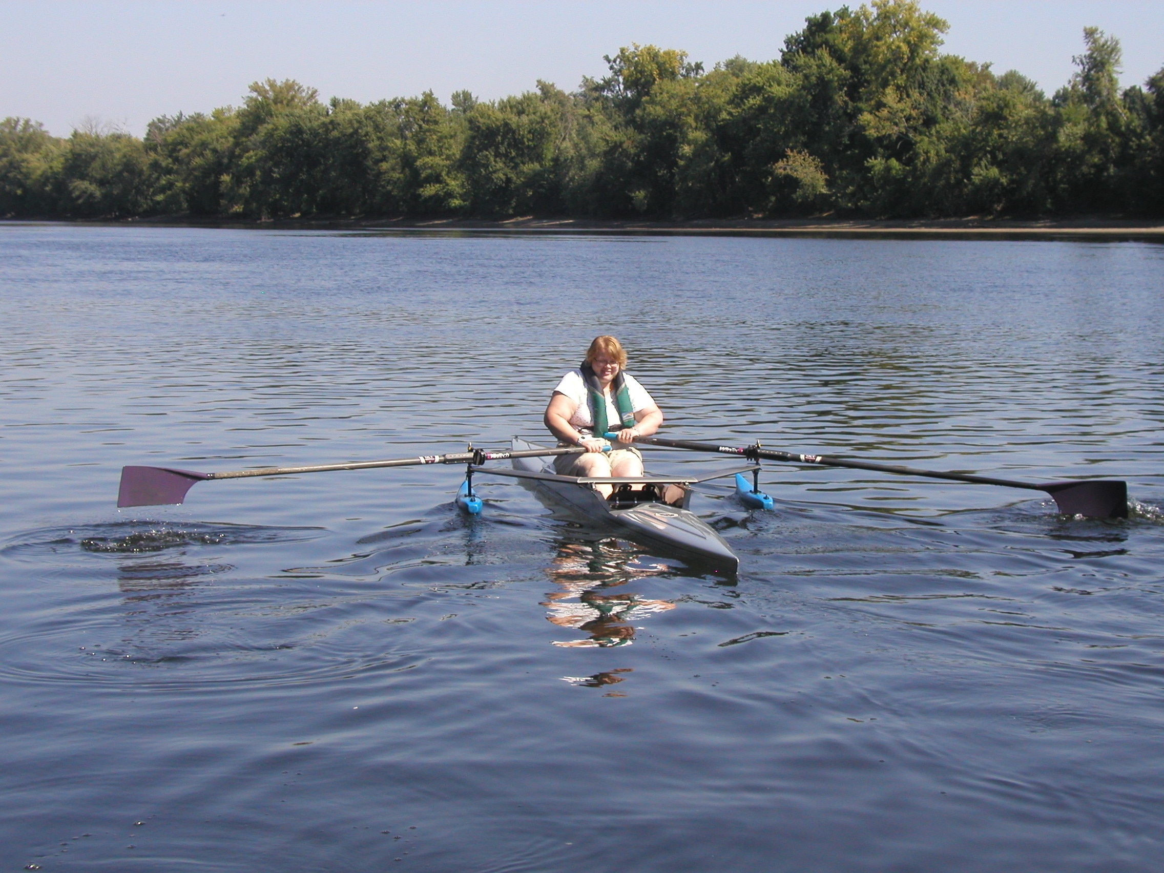 A person wearing a thin lifejacket is sculling in a shell with outriggers on it. 