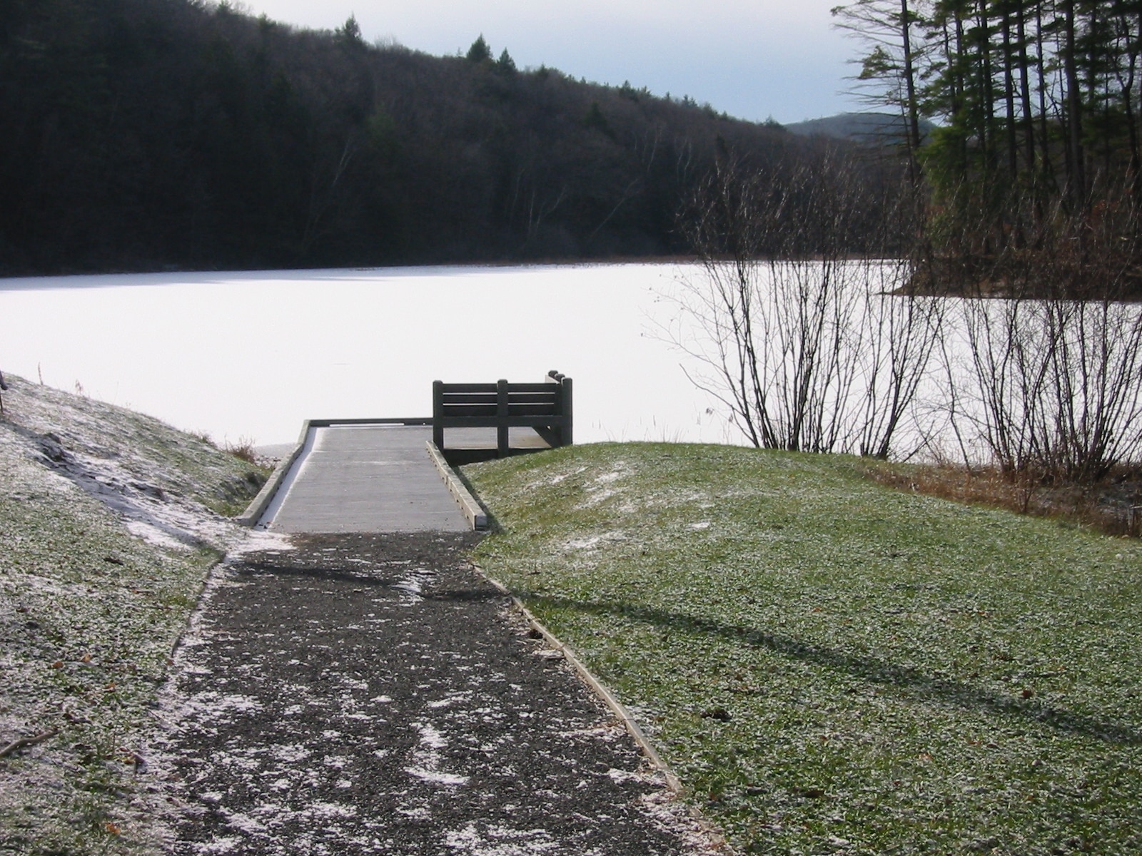 A paved path slopes gently down to a platform with a curb. The platform extends over a lake covered in ice and snow.