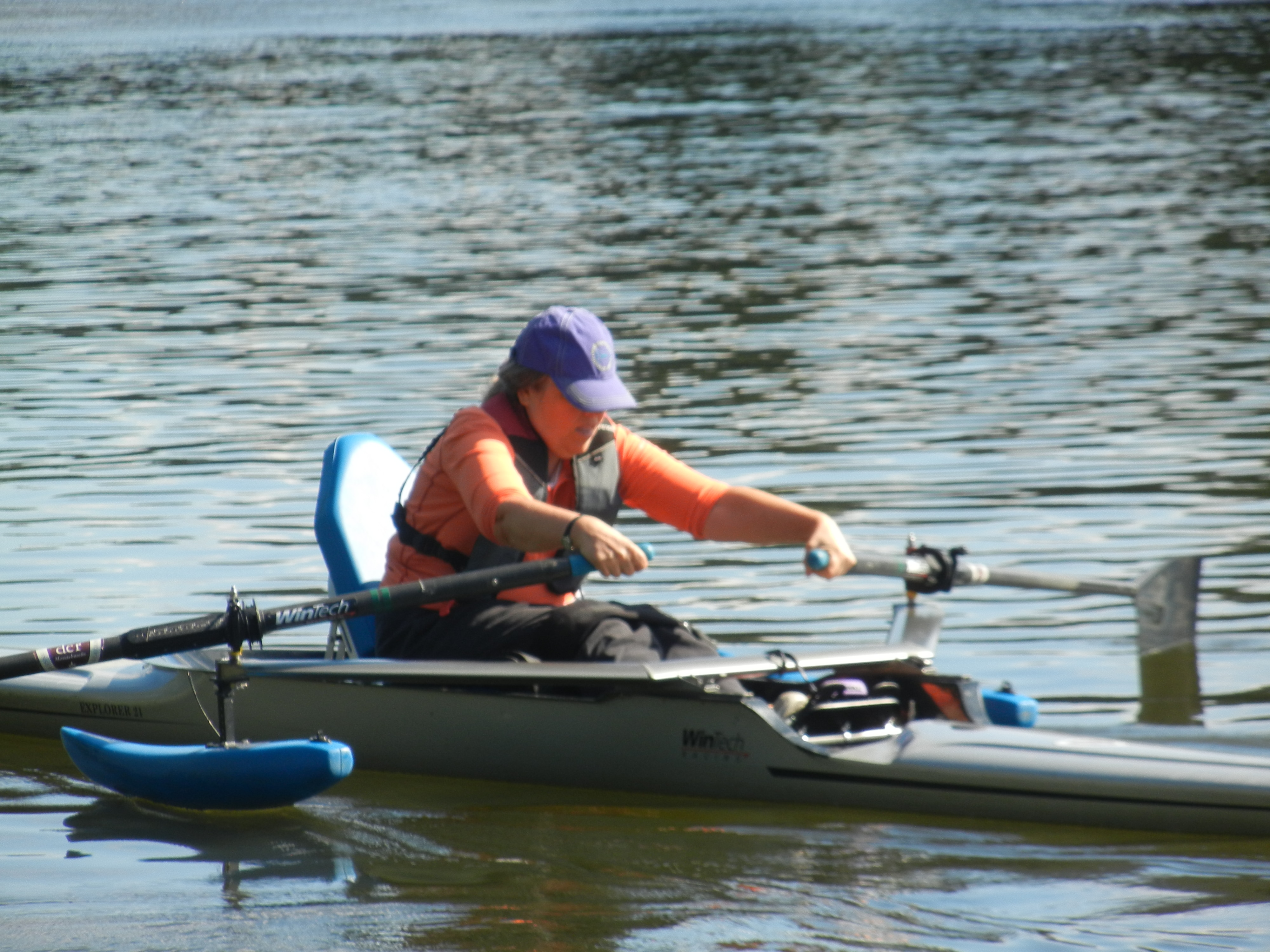 A person is sculling in a shell with pontoons, reaching far forward. The shell has a seat with a high back.