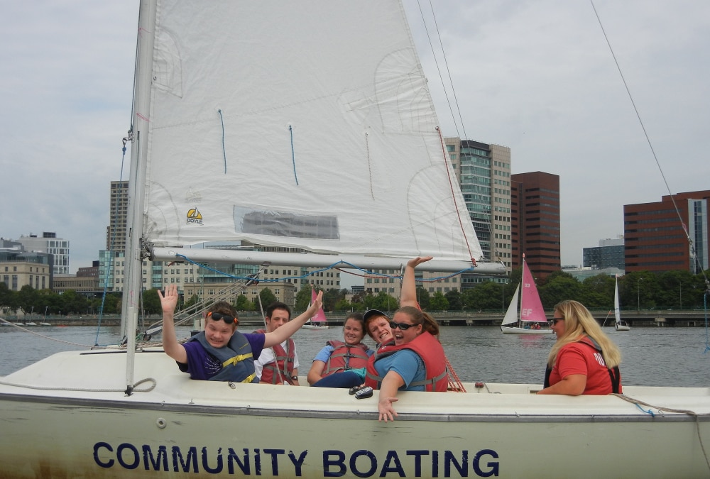 Six people are sharing a sailboat. The people in the front of the boat are smiling and waving, while one person in the back controls the rudder.