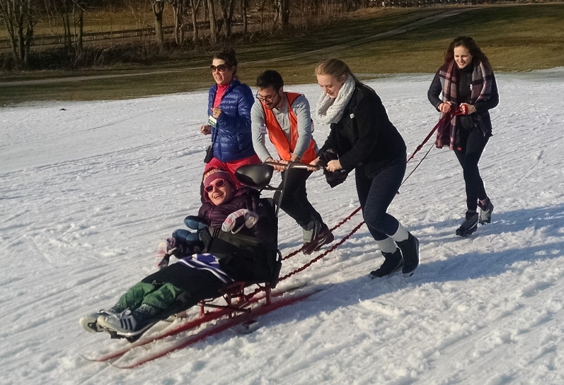 A sit-skier is using a straight-legged ski with handles and outriggers. Two people standing behind the sit ski are pushing on the handle to move the ski up a slight hill. Another person is holding a long rope attached to the back of ski. A fourth person is jogging alongside.