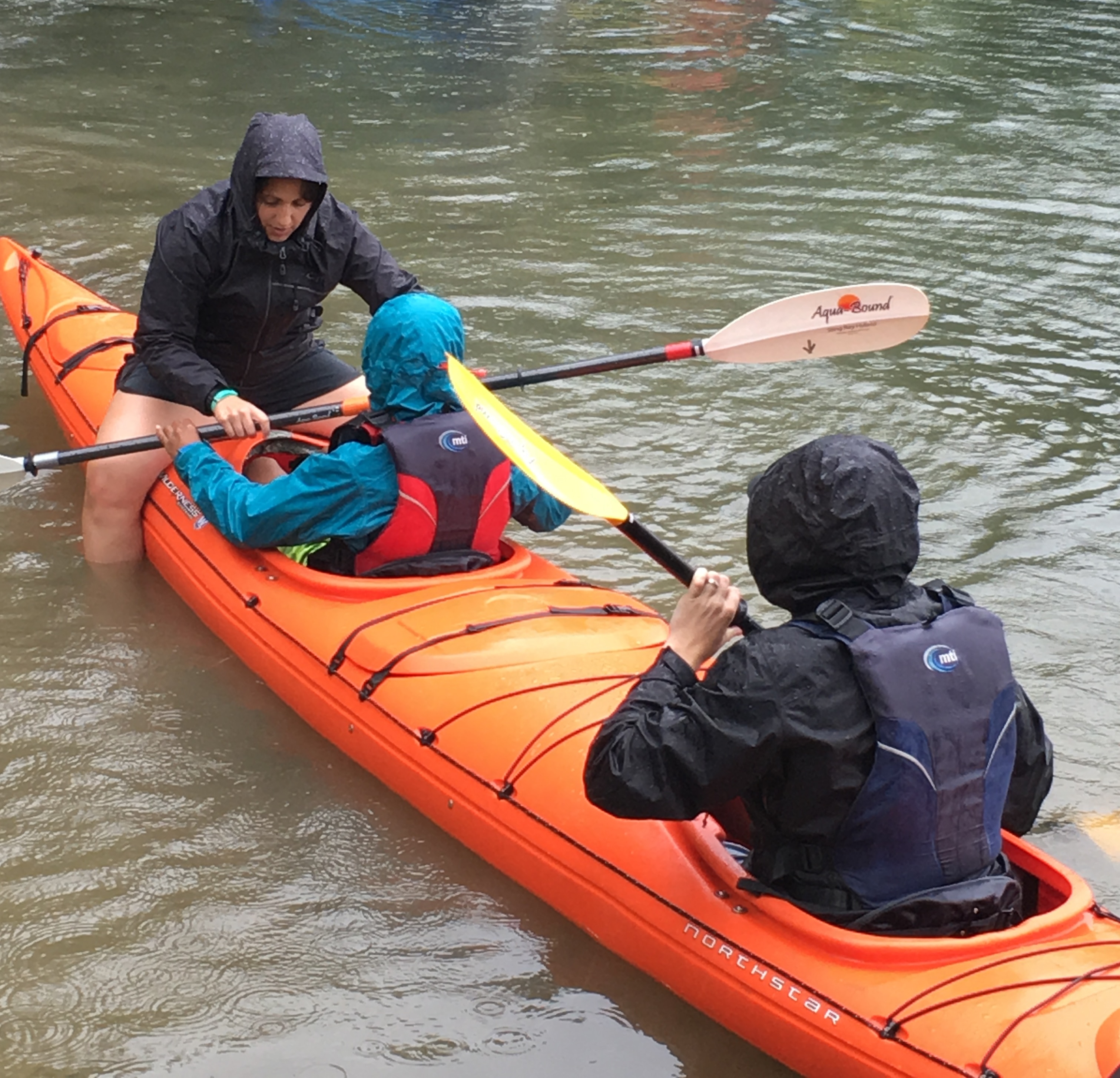 Two paddlers share a tandem kayak with two small cockpits. An instructor is straddling the kayak and giving instructions on paddle use.