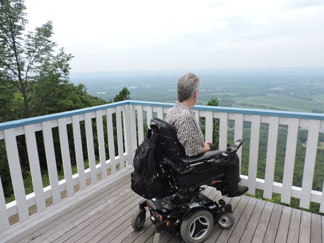 A person using a power wheelchair is looking over the railing on a deck. Fields are visible far below in the distance.